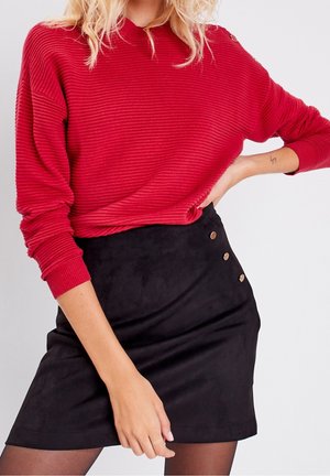 Femme portant un pull rouge côtelé rentré dans une jupe noire à boutons, posant avec une main sur la hanche devant un fond uni.