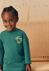 Grøn langærmet skjorte lavet af bomuld med et gult ande-motiv og teksten "QUACKING FUN" på brystet. Blød tekstur, afslappet pasform.