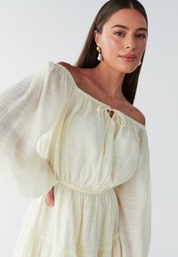 Abito scivolato color crema chiaro, con tessuto strutturato, maniche a sbuffo e vita increspata con dettaglio annodato.