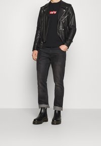 Mężczyzna w czarnej skórzanej kurtce motocyklowej, czarnym t-shircie Levi's, ciemnych wywiniętych dżinsach i czarnych skórzanych butach, stojący na tle białym.