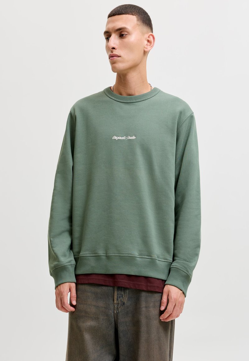 Homme portant un sweat-shirt vert à col rond avec un texte blanc, une chemise rouge foncé en dessous et un pantalon marron ample, regardant sur le côté sur un fond uni.