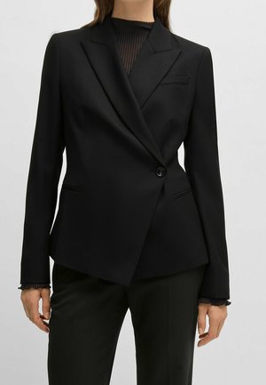 Vrouw draagt een op maat gemaakte zwarte blazer met een enkele knoop, met ingesneden revers, en daaronder een zwart geplooid doorschijnend topje, gecombineerd met een zwarte broek.