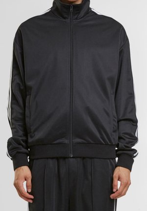 Man wearing a black zip-up track jacket with white stripes on sleeves and matching black pants, standing against plain background.