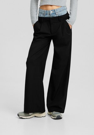 Zwarte, brede broek met een lichtblauwe denim tailleband. Beschikt over voorzakken en een gladde textuur. Gepaard met grijze sneakers.