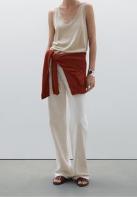 Beżowy bezrękawnik z dekoltem w serek, zestawiony z kremowymi spodniami o szerokich nogawkach i pomarańczowo-czerwonym dzianym swetrem związanym w talii; brązowe płaskie sandały.