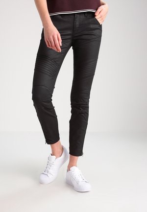 Personne portant un pantalon noir près du corps avec des détails nervurés aux genoux et des baskets blanches, debout avec une main dans la poche et les jambes croisées.
