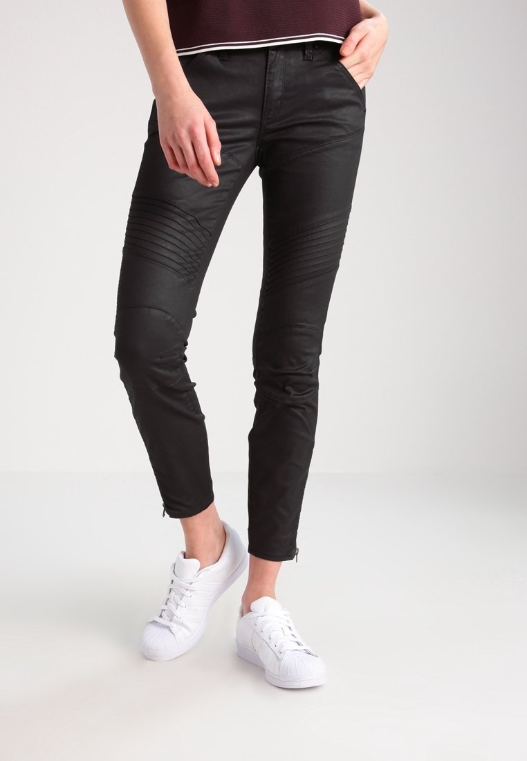 Personne portant un pantalon noir près du corps avec des détails nervurés aux genoux et des baskets blanches, debout avec une main dans la poche et les jambes croisées.