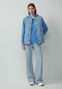 Gilet trapuntato blu con bottoni a pressione, due tasche, indossato sopra un felpa blu. Jeans a gamba larga blu chiaro e sneakers bianche completano l'outfit.
