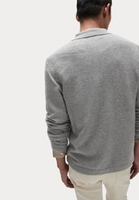 Grå sweater med en blød struktur, lange ærmer og en afslappet pasform. Manchetterne viser et stribet mønster. Båret over lyse bukser.