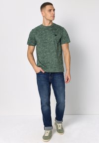 Zielony t-shirt w wzory wykonany z bawełny, zestawiony z niebieskimi dżinsami. Posiada okrągły dekolt i mały czarny detal z logo. Obuwie to zielone sneakersy.