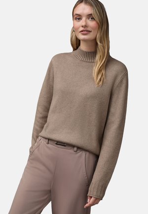 Beżowy sweter dzianinowy z żebrowanym wysokim kołnierzem, długimi rękawami i luźnym krojem, w zestawie z jasnobeżowymi spodniami z kieszeniami bocznymi.