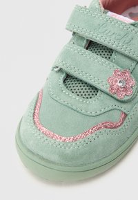 Green suede sneaker with mesh panels, dual Velcro straps, pink glitter accents, and a decorative glitter flower detail.