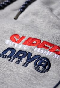 Grijze stof met gedurfde, geborduurde tekst "SUPERDRY" in het rood en "DRYR" in het marineblauw. Accentueert met horizontale lijnen en een zwart veterdetail.