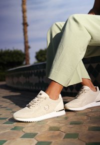 Beige pletené tenisky s texturovaným povrchem, s kovovým logem a kontrastní bílou podrážkou. Nosené s světle zelenými kalhotami.
