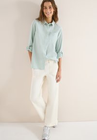 Femme souriante portant une chemise à rayures vert clair à boutons avec les manches retroussées, un pantalon blanc à jambes larges et des baskets argentées, debout de manière décontractée.