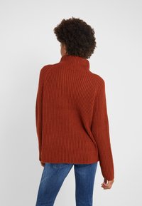 Rust-colored chunky knit sweater with a high collar, ribbed texture, and relaxed fit, paired with blue denim jeans.