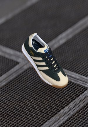En enkelt sort-hvid Adidas-sneaker med sorte snørebånd, der svæver over en metalristet overflade.