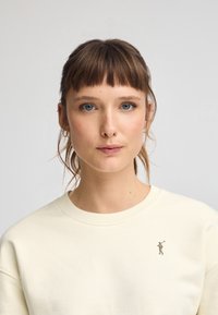 Sudadera beige claro con cuello redondo y puños acanalados, que presenta un pequeño logo bordado en el lado izquierdo del pecho. Textura suave y lisa.