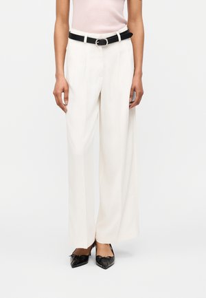 Femme portant un pantalon blanc taille haute à jambes larges, une ceinture noire et des talons pointus noirs ornés de nœuds, debout devant un fond uni.