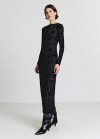 Robe longue noire en tissu transparent, ornée de broderies florales, manches longues et col montant. Associée à des bottines noires à bout pointu.