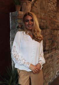 Biela blúzka s čipkovými detailmi na dlhých rukávoch, zapínanie na gombíky vpredu a jemnou textúrou. Nosí sa s béžovými kordovými nohavicami v rustikálnom prostredí.
