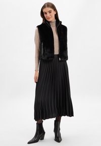 Czarna kamizelka z futra syntetycznego z wysokim kołnierzem i zamkiem błyskawicznym, zestawiona z plisowaną czarną spódnicą, noszona z czarnymi botkami za kostkę.