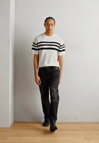 Weißes T-Shirt mit schwarzen horizontalen Streifen, kombiniert mit schwarzen Jeans und schwarzen Schuhen. Modell läuft auf holzverkleideten Boden.