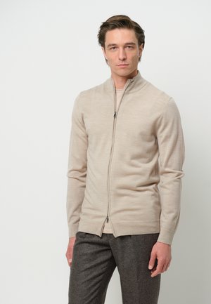 Beige zip-up cardigan made of soft material, featuring a high collar, long sleeves, and a fitted design. Smooth texture, no noticeable patterns.