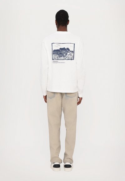 Maglietta bianca a maniche lunghe con un grafico paesaggistico blu sulla schiena, abbinata a pantaloni beige chiaro e sneaker bianche con dettagli neri.