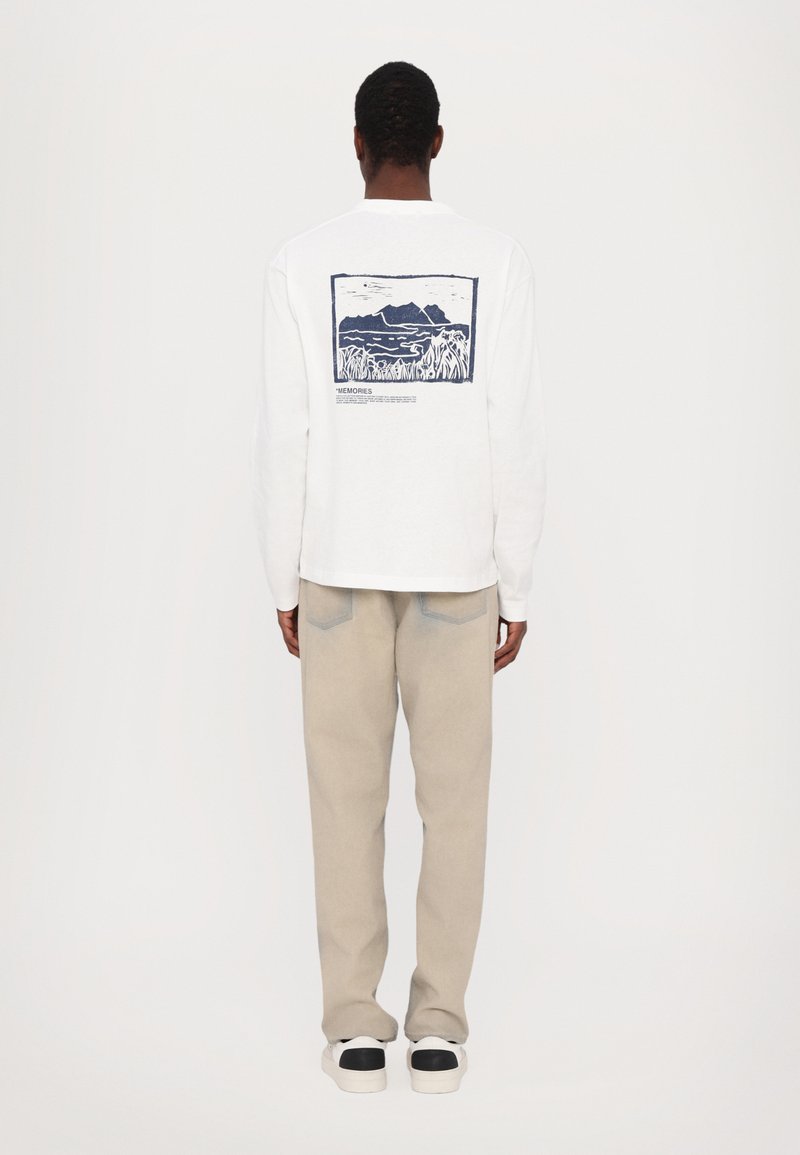 Hvit langermet t-skjorte med en blå landskapsgrafikk på baksiden, kombinert med lysebeige bukser og svarte detaljer på hvite sneakers.