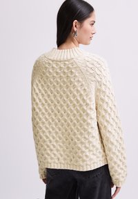 Pull en tricot crème avec un motif en nid d'abeille, un col et des poignets côtelés. Présente une coupe ample et des coutures d'épaule tombantes.