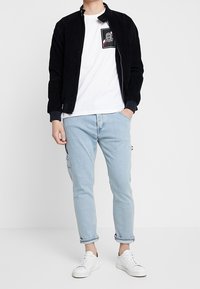 Chaqueta negra de cordón con cremallera, camiseta blanca con un logo, jeans ajustados de color azul claro y zapatillas blancas. Diseño minimalista con corte holgado.