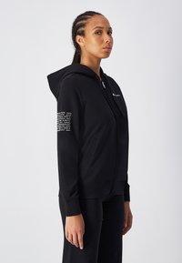 Sudadera negra con cremallera y capucha ajustable, que presenta un patrón texturizado en la manga derecha y un logo en el frontal. Tela suave, puños acanalados.