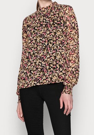 Frau trägt eine transparente, langärmelige Bluse mit Blumenmuster in Pink und Beige auf Schwarz, kombiniert mit schwarzer Hose vor schlichtem Hintergrund.