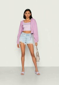 Mulher vestindo um hoodie curto lilás, uma blusa xadrez curta, calções de ganga rasgados, sandálias lilás e segurando uma bolsa bege contra um fundo liso.