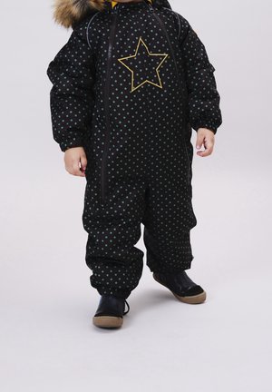 Mono de nieve acolchado negro con estampado de estrellas verdes y una estrella amarilla de acento. Cuenta con capucha con ribete de piel y puños elásticos. Combinado con botas negras.