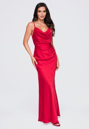 Femme portant une robe de soirée rouge en satin sans manches avec un décolleté bénitier et une taille ajustée, debout devant un fond uni gris clair.