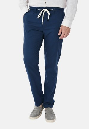 Uomo che indossa pantaloni blu navy con coulisse bianca, camicia bianca a maniche lunghe e scarpe casual grigie senza lacci, in piedi con le mani in tasca.