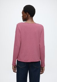 Gari piedurkņu krekls gaiši rozā krāsā, izgatavots no teksturēta auduma. Raksturots ar vaļīgu piegriezumu un apaļu kakla izgriezumu, kombinēts ar tumšām džinsa biksēm.