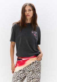T-shirt en coton gris à manches courtes avec un logo imprimé, associé à un tissu abstrait multicolore drapé sur un pantalon à imprimé léopard.