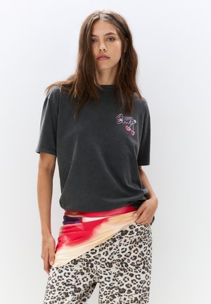 T-shirt en coton gris à manches courtes avec un logo imprimé, associé à un tissu abstrait multicolore drapé sur un pantalon à imprimé léopard.