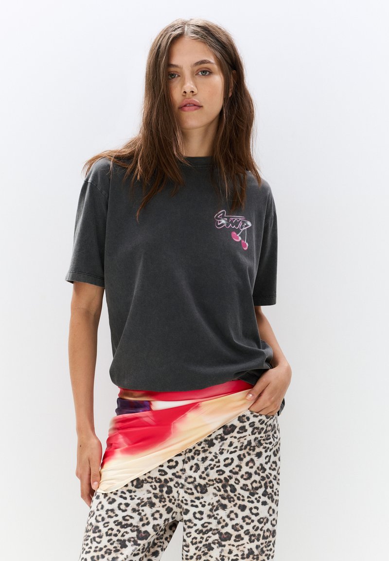 T-shirt en coton gris à manches courtes avec un logo imprimé, associé à un tissu abstrait multicolore drapé sur un pantalon à imprimé léopard.
