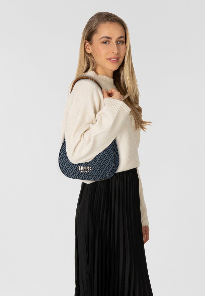 Sac à main à motif bleu avec une forme incurvée, doté d'une bandoulière et de ferrures dorées. Porté avec un pull crème et une jupe plissée noire.