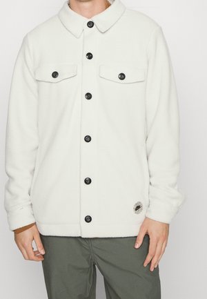 Veste en polaire blanche avec une fermeture à boutons sur le devant, boutons noirs, deux poches poitrine, un col rabattable et un patch logo sur le bas.