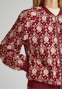 Bordeaux bomberjack met een geometrisch patroon in crème- en gele tinten, met ribgebreide manchetten en een ritssluiting. Glad textieloppervlak.