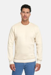 Sudadera de color crema con cuello redondo acanalado y acentos a rayas en los puños y el dobladillo, combinada con jeans azules.