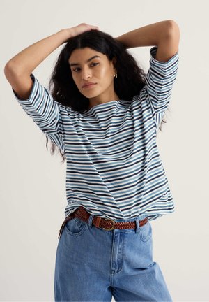 Femeie cu păr creț și întunecat, purtând o cămașă cu dungi albastre și albe și blugi albaștri, pozând cu mâinile după cap pe un fundal simplu.