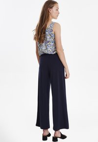 Ärmelloses Oberteil mit floralem Muster in Blau, kombiniert mit weiten, lockeren navy-blauen Hosen. Das Modell trägt schwarze Slipper. Rückansicht präsentiert.