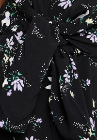 Vestido de tela negra con un estampado floral en púrpura y blanco, con acentos verdes. Hay un detalle de diseño anudado en la cintura.