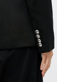 Musta räätälöity bleiseri, jossa on neljä hopeista nappia logokoristeilla hihassa, teksturoitu kangas ja tyylikäs muoto.